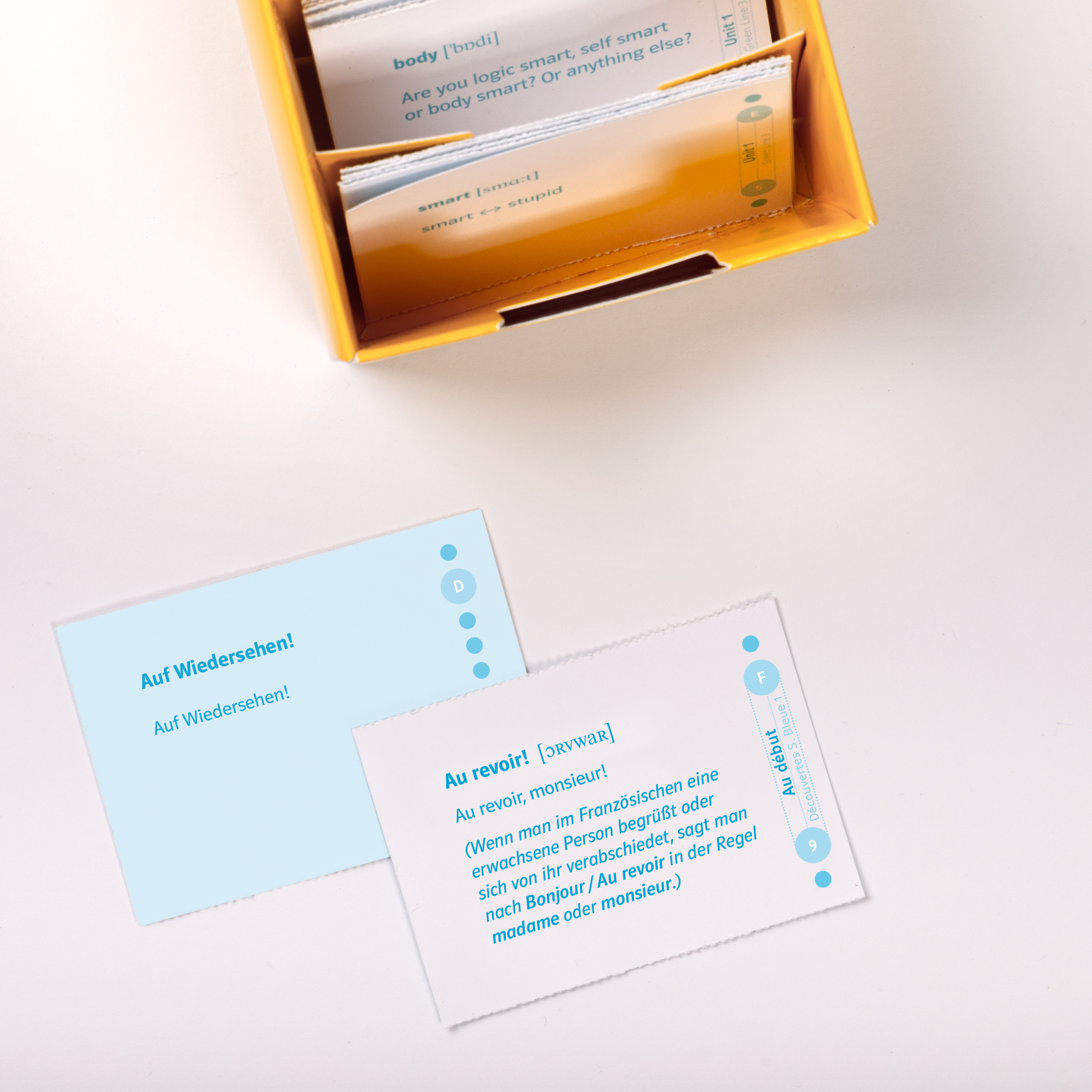 Découvertes 1 Série bleue - Vokabel-Lernbox zum Schülerbuch Eine gelbe Schachtel enthält Sprachlernkarten, wobei zwei Karten auf einer weißen Fläche die Sätze Auf Wiedersehen! auf Deutsch und Au revoir! auf Französisch abbilden.